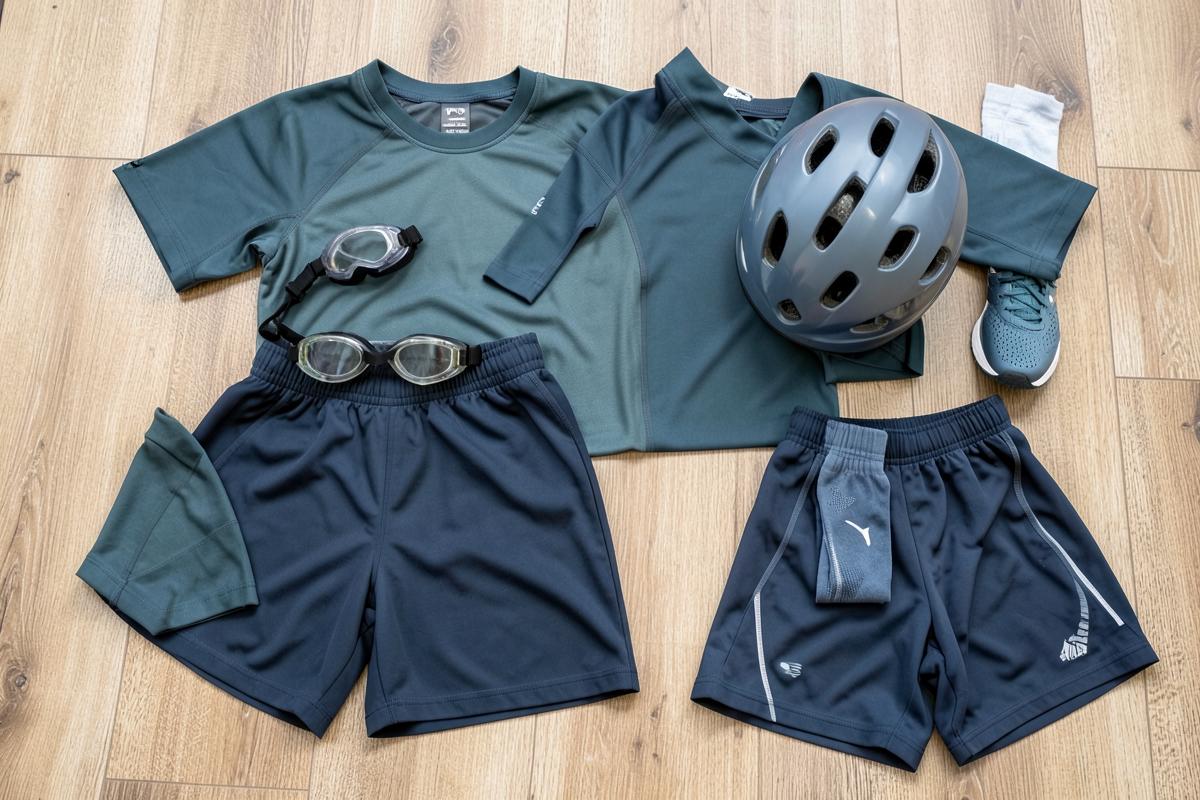 Basic beginner triathlon kit laid out on a floor: goggles, swim shorts, helmet, running shoes, socks and sports clothing
