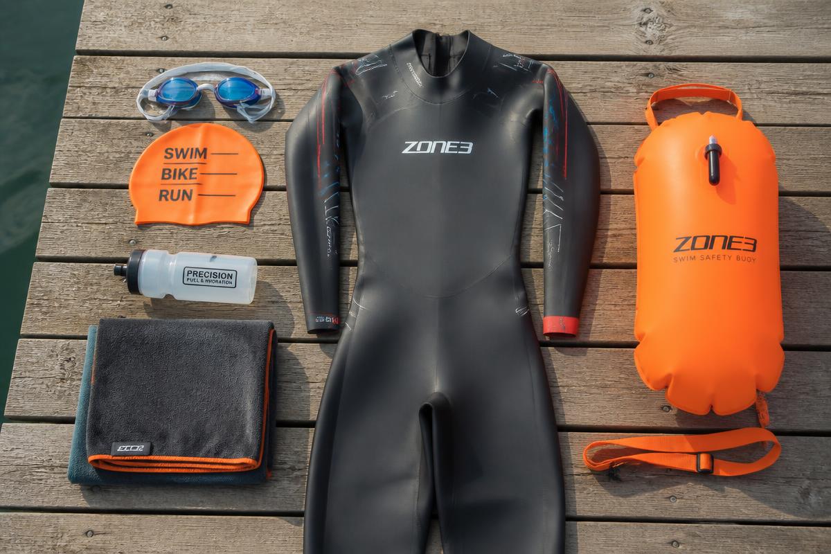 Open water swim kit laid out on a jetty: goggles, bright orange cap, wetsuit, safety buoy, towel and water bottle