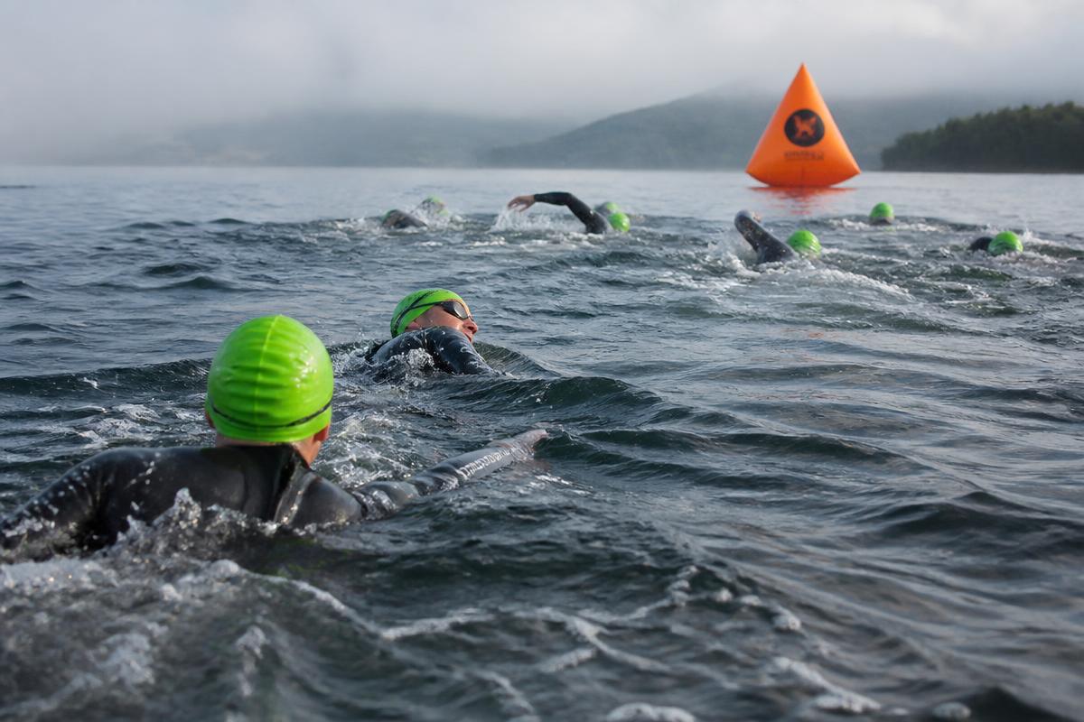 Open water swimmers in wetsuits and bright caps swimming towards a large orange turn buoy on a misty lake