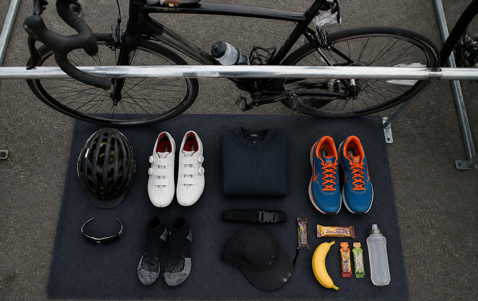 Helmet, cycling shoes, running shoes, sunglasses and nutrition laid out neatly beside a racked triathlon bike