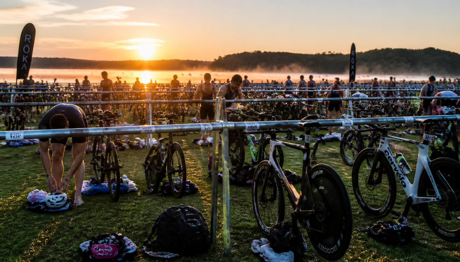 Triathlon transition area at sunrise, bikes racked in rows with athletes preparing kit beside a misty lake