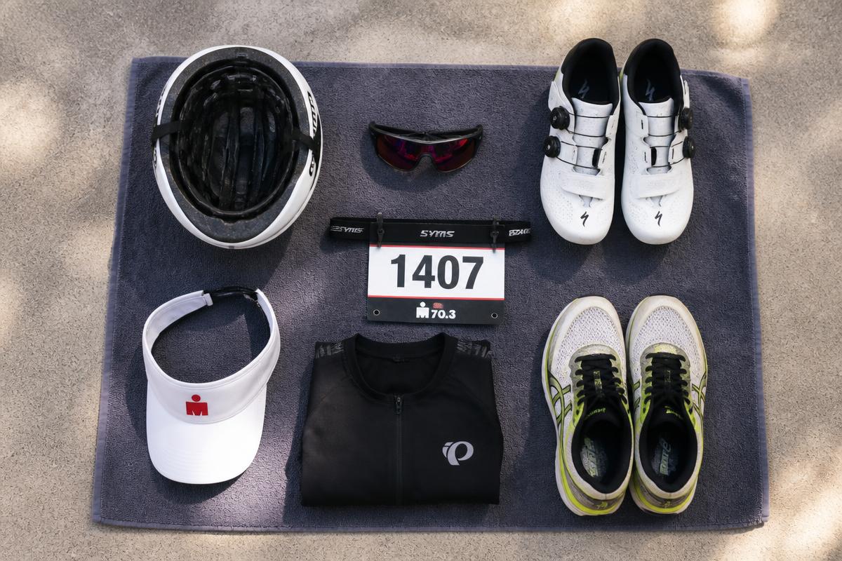 Flat-lay of triathlon transition kit including helmet, sunglasses, cycling shoes, running shoes, race belt and number on a towel